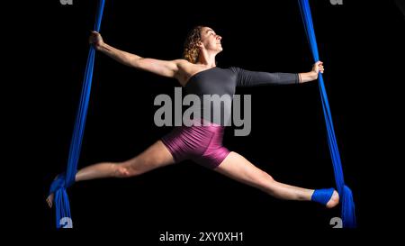 Femme s'engage dans une danse aérienne élégante utilisant des soies bleues vibrantes, montrant la flexibilité et la force dans une pose partagée dynamique, isolée contre un bla Banque D'Images