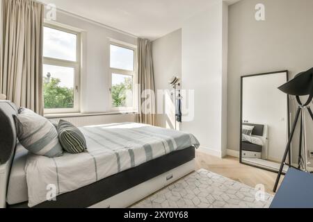 Cette chambre moderne lumineuse et aérée comprend un grand lit avec du linge de maison rayé, des fenêtres avec des rideaux et un mobilier minimaliste. Banque D'Images