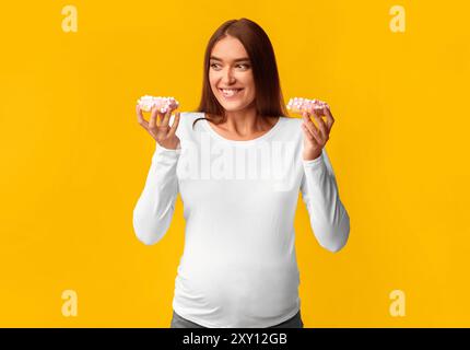 Fille enceinte Holding Donuts debout sur fond rose, Studio Shot Banque D'Images