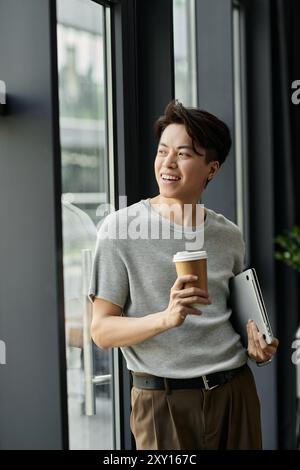 Un jeune homme tient une tasse de café et regarde par une fenêtre dans un immeuble de bureaux. Banque D'Images