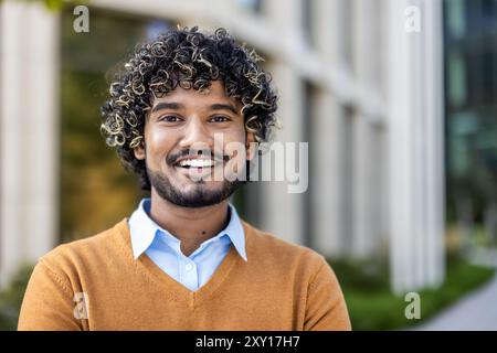 Homme d'affaires indien confiant avec les cheveux bouclés souriant à l'extérieur dans l'environnement urbain. Portant un pull orange et une chemise bleue, représentant un style professionnel mais décontracté. Banque D'Images