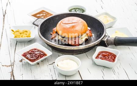 Hamburger dans une poêle à frire avec des sauces placées autour sur fond de bois blanc avec de la peinture fissurée. Banque D'Images