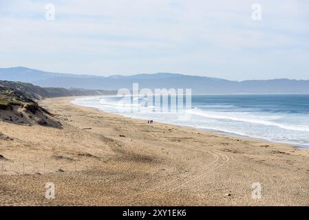 Les gens marchant le long d'une grande plage de sable bordée de hautes dunes de sable un jour d'automne brumeux. D'énormes vagues se brisant sur la plage sont visibles à distance. M Banque D'Images