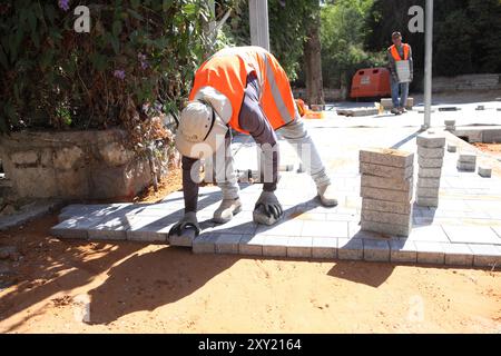 Travailleur palestinien avec gilet de protection met des briques sur des couches de béton comprimé et de sol de sable un autre apporte plus de briques, ils reconstruisent un trottoir. Banque D'Images