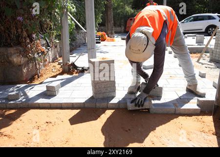 Travailleur palestinien avec gilet de protection met des briques sur des couches de béton comprimé et de sol de sable, il travaille à la reconstruction d'un trottoir. Banque D'Images