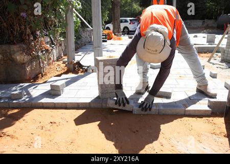 Travailleur palestinien avec gilet de protection met des briques sur des couches de béton comprimé et de sol de sable, il travaille à la reconstruction d'un trottoir. Banque D'Images