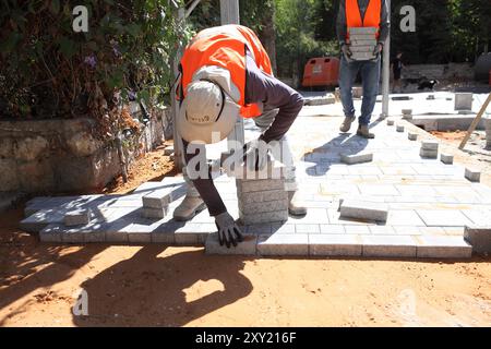 Travailleur palestinien avec gilet de protection met des briques sur des couches de béton comprimé et de sol de sable un autre apporte plus de briques, ils reconstruisent un trottoir. Banque D'Images