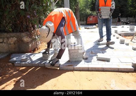 Travailleur palestinien avec gilet de protection met des briques sur des couches de béton comprimé et de sol de sable un autre apporte plus de briques, ils reconstruisent un trottoir. Banque D'Images