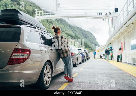 Femme s'appuie contre sa voiture sur le ferry norvégien entouré par d'autres véhicules avec des montagnes vertes brumeuses en arrière-plan. Scène capture l'essence de relaxe Banque D'Images