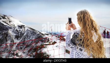 Blonde woman photographing paysage d'hiver montagnes et neige avec cell phone Banque D'Images
