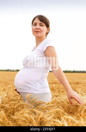 Femme enceinte en T-shirt blanc touchant le ventre et regardant la caméra tout en se tenant debout dans le champ de seigle à la campagne Banque D'Images