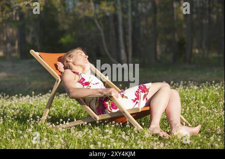 Une jeune femme dans une robe d'été se détache dans un transat classique et prend des bains de soleil. Le transat est debout dans une prairie pleine de girofle fraîche fleurie Banque D'Images