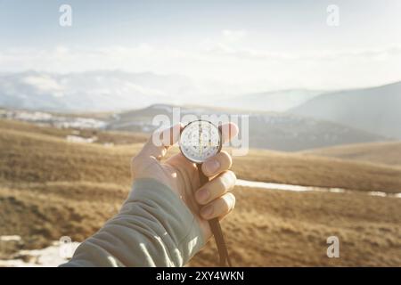 La main d'un homme tient une boussole à main sur fond de montagnes et de collines au coucher du soleil. Le concept de voyage et de navigation dans les espaces ouverts Banque D'Images