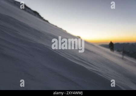 L'alpiniste floue équipé professionnellement à l'aube du soleil marche sur la pente tôt le matin et la neige dérive sur la neige au-dessus de la Banque D'Images
