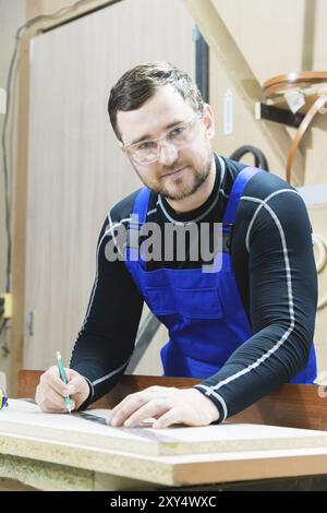 Un beau charpentier fatigué barbu sur une table avec un crayon dessine un signe sur le tableau. Sur le lieu de travail, il y a un maître ordinaire habillé en bleu CA Banque D'Images