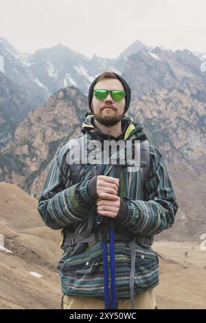 Un gars avec une barbe et portant des lunettes de soleil dans une veste à membrane, chapeau, avec un sac à dos et des bâtons pour la marche nordique, un voyageur debout dans l'ai ouvert Banque D'Images