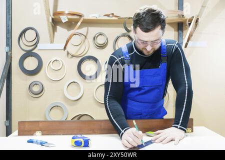 Un beau charpentier fatigué barbu sur une table avec un crayon dessine un signe sur le tableau. Sur le lieu de travail, il y a un maître ordinaire habillé en bleu CA Banque D'Images