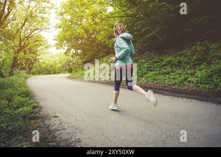 Jeune femme blonde de forme physique dans un casque courant le matin sur la forêt caucasienne sentier dans la lumière du soleil. Vue latérale de derrière Banque D'Images