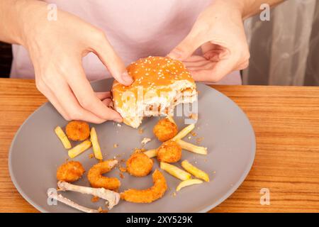 Un homme caucasien mange des hamburgers à moitié mangés, des chips de pommes de terre, des rondelles d'oignon et des morceaux de poulet à la main dans une assiette grise sur une table en bois Banque D'Images