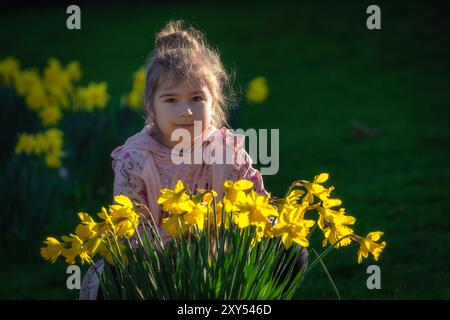 Jeune fille assise derrière des fleurs de jonquille jaunes illuminées par la lumière du soleil, souriant et regardant la caméra à Phoenix Park, Dublin, Irlande Banque D'Images