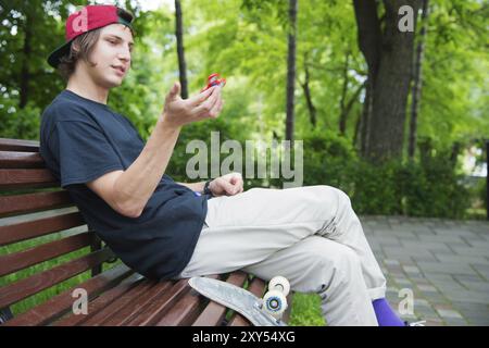 Un patineur hipster aux cheveux longs dans une casquette est assis sur un banc et tourne une spineless spinner à côté d'une planche à roulettes Banque D'Images