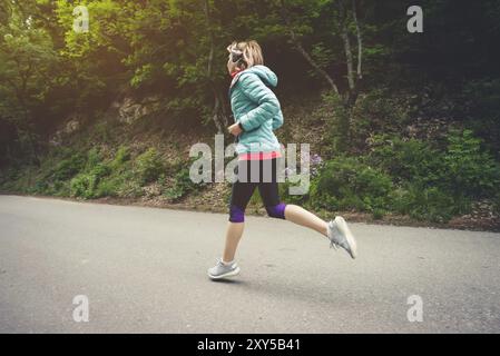 Jeune femme blonde de forme physique dans un casque courant le matin sur la forêt caucasienne sentier dans la lumière du soleil. Vue latérale de derrière Banque D'Images