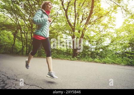 Jeune femme blonde de forme physique dans un casque courant le matin sentier de forêt caucasienne à la lumière du soleil Banque D'Images