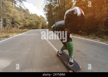 Un jeune cavalier dans une combinaison spéciale en cuir et un casque pleine longueur roule à grande vitesse sur son longboard sur une route de campagne dans la forêt d'automne Banque D'Images