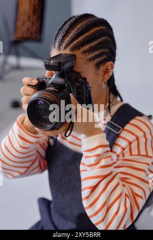 Portrait vertical d'une jeune femme afro-américaine prenant des photos en studio à l'aide d'un appareil photo reflex numérique professionnel et regardant dans le viseur Banque D'Images