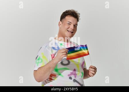 Une personne queer sourit largement, tenant un drapeau arc-en-ciel dans une main. Banque D'Images