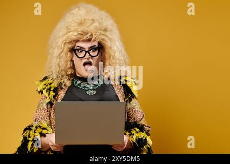 Une drag queen dans une tenue à la mode et une grande perruque réagit à quelque chose sur leur ordinateur portable. Banque D'Images