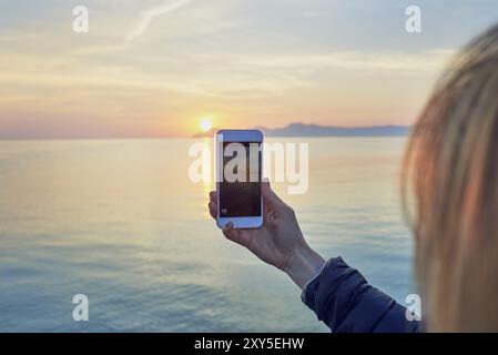 Jeune femme blonde prenant une photo d'un coucher de soleil coloré sur l'océan sur un téléphone portable avec une vue rapprochée de l'écran du téléphone portable avec la mer et le ciel Banque D'Images