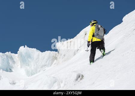 Un skieur dans un casque et un masque avec un sac à dos s'élève sur une pente sur fond de neige et un glacier avec piolet à la main. Backcountry Fre Banque D'Images