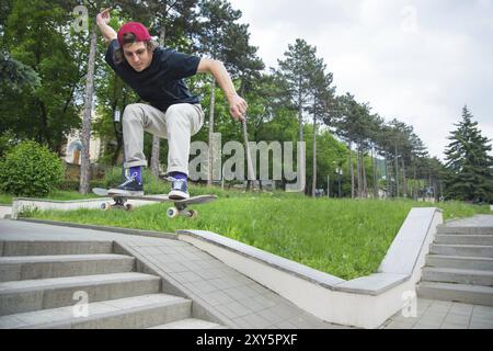 Patineur-adolescent aux cheveux longs dans un T-shirt et casquette de baskets saute l'ollie sur fond d'un ciel orageux à travers la ville Banque D'Images