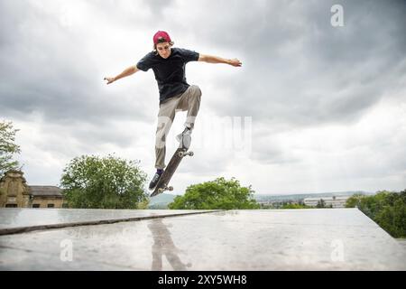 Patineur-adolescent aux cheveux longs dans un T-shirt et casquette de baskets saute l'ollie sur fond d'un ciel orageux à travers la ville Banque D'Images