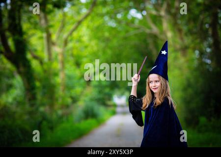 Fille d'enfant blonde caucasienne en costume bleu sorcier avec cape et chapeau, jetant un sort avec baguette magique dans un fond de forêt extérieure. Banque D'Images