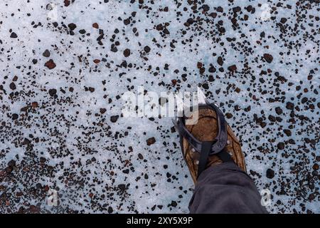 Chaussure de randonnée avec des crampons sur la glace avec des pierres volcaniques autour, vue de dessus, glacier Skaftafell dans le parc national de Vatnajokull, Islande. Randonnée glaciaire. Banque D'Images