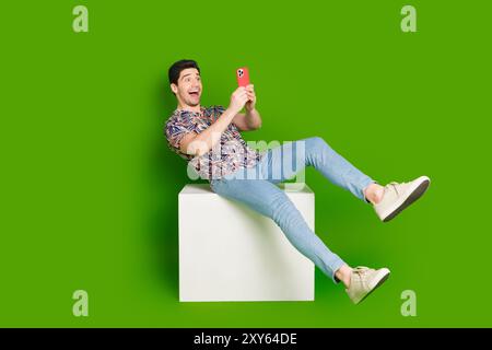 Photo pleine taille de jeans de chemise imprimée habillée par un gars surjoyeux assis sur un cube blanc regarder un smartphone isolé sur fond de couleur verte Banque D'Images