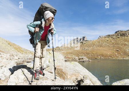 Randonneur fille mince et sympathique dans des lunettes de soleil et une veste en duvet mince bleue avec un sac à dos et des bâtons de trekking s'élève à un haut rocher contre le backgro Banque D'Images