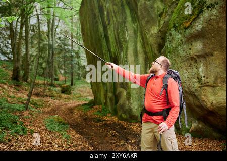 Les points de randonneur avec le bâton de trekking vers la formation rocheuse imposante dans la forêt luxuriante. Portant un grand sac à dos, stands touristiques sur un sentier couvert de feuilles entouré de verdure verdoyante. Exploration et découverte. Banque D'Images