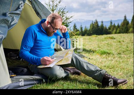 L'homme est assis dans l'entrée de la tente touristique, profondément concentré sur la carte. Touriste tient la boussole et contemple son itinéraire, entouré d'un champ verdoyant luxuriant avec des arbres et des montagnes à distance sous le ciel nuageux. Banque D'Images