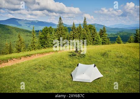 Tente touristique blanche au soleil, au sommet d'une colline herbeuse, entourée de jeunes pins. Vue panoramique sur les montagnes luxuriantes et vallonnées sous un ciel bleu vif avec des nuages dispersés, créant un endroit de camping idyllique. Banque D'Images