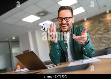 Vue en angle bas d'un chef de projet joyeux envoyant des messages vocaux sur son téléphone portable sur son lieu de travail Banque D'Images