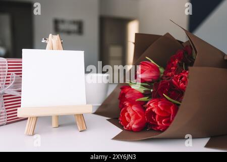 Bouquete de tulipes rouges sur table, tasse à café, cadre photo blanc vide. Concept de carte de vœux. Fête des mères, fête internationale des femmes ou fête de la Saint-Valentin Banque D'Images