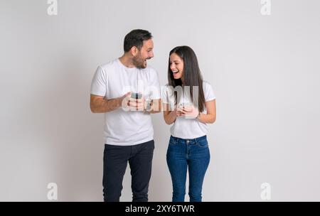 Jeune petit ami et petite amie avec des téléphones intelligents texto et riant joyeusement sur fond blanc Banque D'Images
