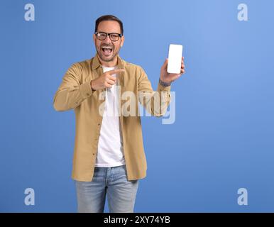 Portrait d'un jeune homme joyeux pointant vers le smartphone et l'application de rencontre publicitaire sur fond bleu Banque D'Images