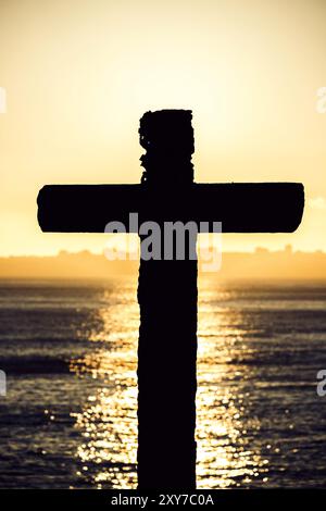 Une croix en bois se dresse sur un coucher de soleil vibrant, avec la lumière dorée reflétant les eaux calmes de l'océan, créant une atmosphère paisible Banque D'Images