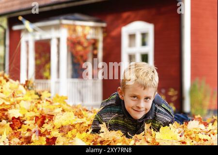 Un garçon allongé dans un grand tas de feuilles d'automne. En arrière-plan une jolie maison suédoise traditionnelle. Un garçon dans un gros tas de feuilles d'automne couché. Dans le Banque D'Images