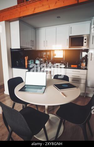 Un intérieur de cuisine moderne avec des armoires blanches, des comptoirs noirs et des appareils électroménagers en acier inoxydable. Une table ronde avec un écran de maquette d'ordinateur portable, ordinateur portable, Banque D'Images