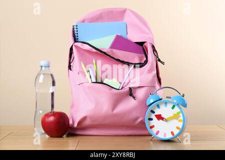 Sac à dos avec différents articles de papeterie scolaire, réveil, bouteille d'eau et pomme sur table en bois sur fond beige Banque D'Images